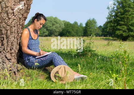 Young indian woman sitting against tree trunk writing in pasture Stock Photo