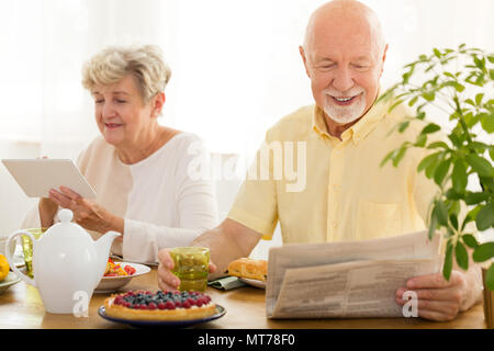 Happy elderly woman using a tablet and her husband reading a newspaper Stock Photo