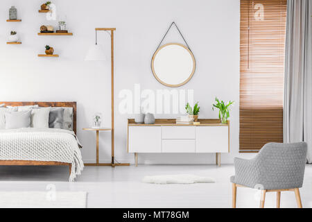 Scandinavian style white room interior with round mirror on the wall, wooden bed with pillows and blanket, cabinet and armchair. Real photo. Stock Photo