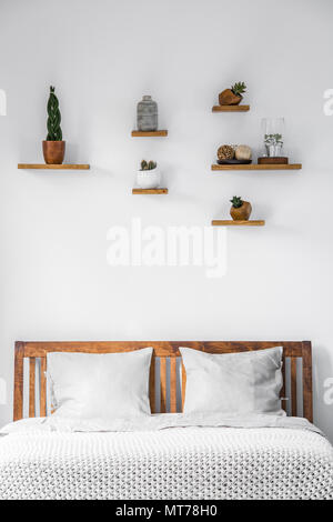 Close-up of a white wall with small decorations on shelves above a wooden headboard of a bed with pillows and blanket in a bright bedroom interior. Re Stock Photo