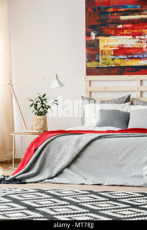Painting above bed with red and grey sheets in bedroom interior with patterned carpet. Real photo Stock Photo