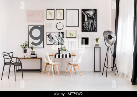 Lamp near white chairs at wooden table in dining room interior with posters and black armchair Stock Photo