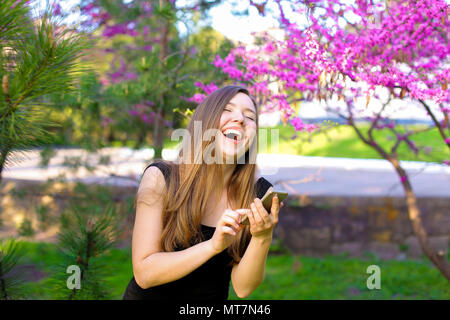 Jocund girl using new smartphone in park with blooming trees background. Stock Photo