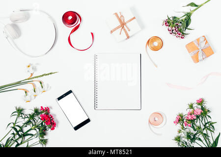 Blank notepad lays on white table near smart phone with empty screen and headphones among flowers, gift boxes and ribbons, top view. Styled desk flat  Stock Photo