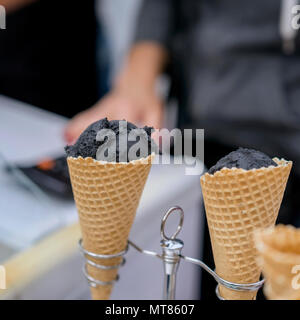 Two waffle cones, horns with balls of trendy black ice cream and hand of the seller. Delicious cooling portion for fun. Selective focus. Real scene in store. Stock Photo