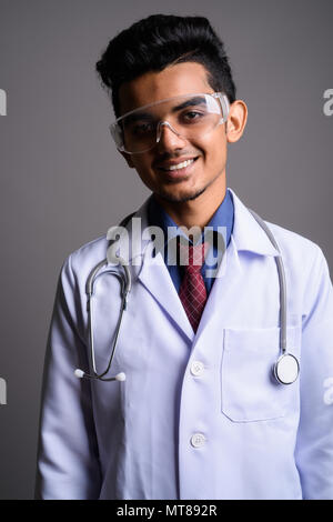 Young Indian man doctor wearing protective glasses against gray  Stock Photo