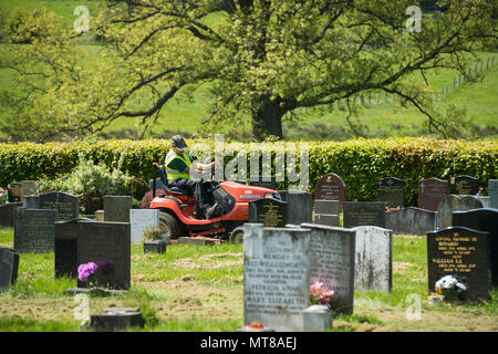 Man working (empoyee of local council) sits on ride on lawn mower & cuts grass between headstones - Guiseley Cemetery, West Yorkshire, England, UK. Stock Photo