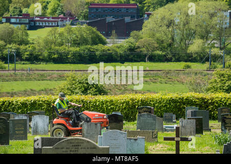 Man working (empoyee of local council) sits on ride on lawn mower & cuts grass between headstones - Guiseley Cemetery, West Yorkshire, England, UK. Stock Photo