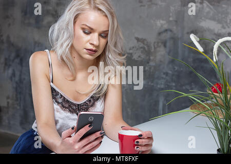 Beautiful blonde girl in home clothes is using a smartphone and smiling while sitting in kitchen Stock Photo