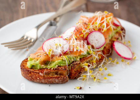 Rustic bread toast with mashed avocado, smoked salmon, radish, broccoli sprouts and seeds served in a white plate on a rustic wooden table. Stock Photo