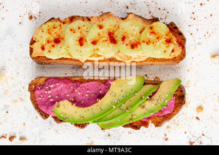 Two healthy vegan toasts with hummus, cucumber and paprika and beetroot hummus, avocado and seeds surrounded by crumbs and seeds on a rustic backgroun Stock Photo