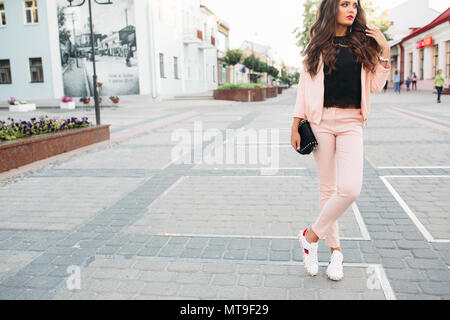 Stylish girl in pink sport chic suit, lace top and sneakers posing in the street with small crossbody bag in hand. Stock Photo