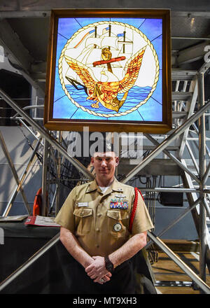 GREAT LAKES, Ill. (Feb. 27, 2018) Builder 1st Class Jonathan Olds stands in  front of a 400-piece stained glass window replica of the Navy Seal, hanging  below the brow of the USS