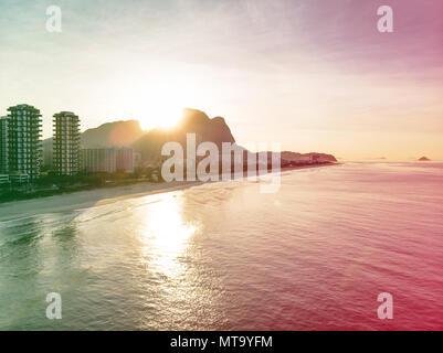 Drone flying towards Barra da Tijuca beach while filming waves crashing on beach during sunrise, with the buildings and the Gavea Stone mountain in si Stock Photo