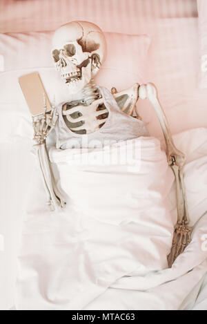 Human skeleton with mobile phone in hand lies in bed, black humor, funny joke Stock Photo