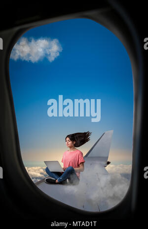 PC addiction, woman with laptop sitting on plane wing, view from airplane porthole Stock Photo