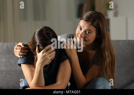 Bad teen is happy with her sad friend crying sitting on a couch in the living room at home Stock Photo