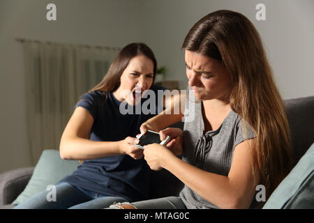 Two friends or sisters fighting for a smart phone sitting on a couch in the living room at home Stock Photo