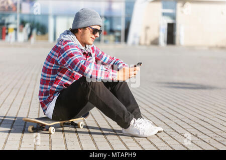 Portrait of hipster man with sunglasses holding smartphone sitting on the skateboard Stock Photo