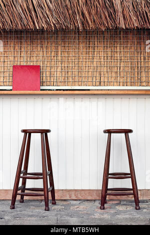 Outdoor cafe bar with two brown wooden stools, an empty red sign, white wooden counter and bamboo cover in an holiday place. Stock Photo
