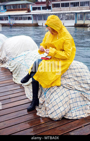 Caucasian woman wearing a yellow raincoat rolling a cigarette on the harbor on a rainy day. She is sitting on the fishing nets near the sea. Stock Photo