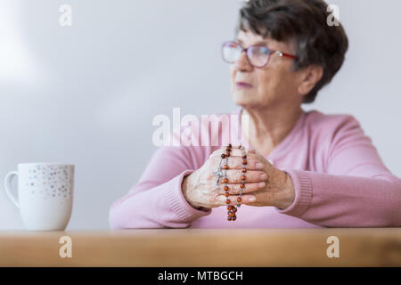 Photo with blurred background of elder woman holding a rosary and sitting by the table with tea mug Stock Photo
