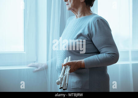 Senior, blind woman holding white cane, standing beside window Stock Photo