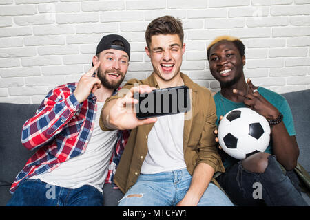 Portrait of three happy young men taking a selfie on phone while sitting at home with beer and snacks indoors Stock Photo