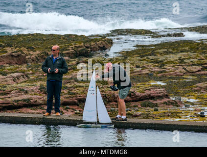 Milsey Bay, North Berwick. East Lothian, Scotland, United Kingdom, 30th May 2018. Men having fun playing with radio controlled Kyosho Marine Racing sailing yacht in the tidal bathing pool with the sea and a wave breaking in the background Stock Photo