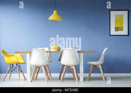 Minimal, modern dining room in interior with a table and contrasting white and yellow chairs around it, against blue wall with a mockup poster Stock Photo