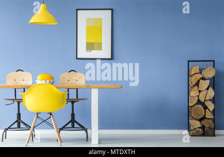 A yellow chair and a hanging lamp in an open space, minimal style interior with a melon in a dish on a table with a pile of firewood against a blue wa Stock Photo