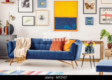 Real photo of a navy blue couch with a blanket and orange pillows standing against a white wall with gallery of posters in living room interior Stock Photo