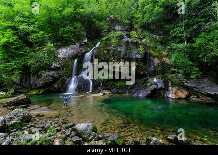 Man in blue jacket standing in shallow water at Virje waterfal with raised hands, Soca valley, Bovec, Slovenia Stock Photo