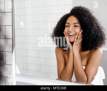 Happy woman admiring her skin in front of the bathroom mirror, touching face with hands Stock Photo