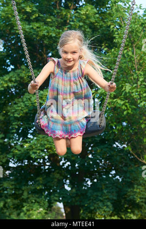 Little child blond girl having fun on a swing outdoor. Summer playground. Girl swinging high. Young child on swing outdoors. Stock Photo