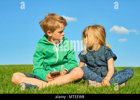 Happy blond children boy and girl using smartphone (watching movie or playing game) sitting on the green grass lawn. Brother and sister. Summertime. Stock Photo