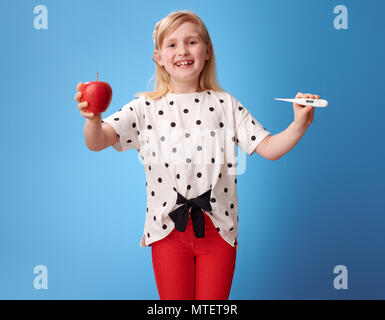 smiling modern child in red pants showing an apple and a thermometer on blue background Stock Photo