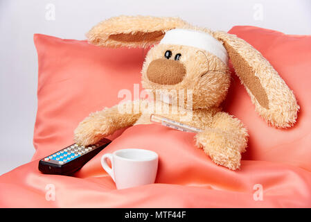 A toy unhealthy rabbit with a thermometer and a TV remote control is lying in bed. Stock Photo