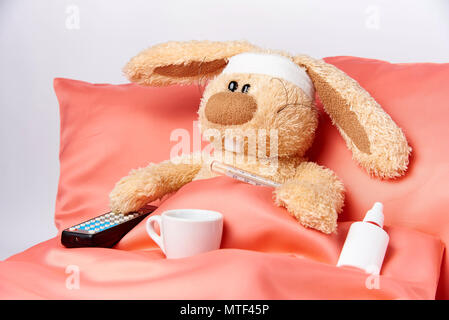 A toy unhealthy rabbit with a thermometer and a TV remote control is lying in bed. Stock Photo