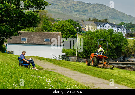 County Council employee cuts the grass whilst a man reads on a bench on a sunny day in Schull, West Cork, Ireland. Stock Photo