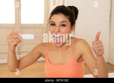 Young beautiful latin woman looking at pregnancy test feeling happy and excited at the positive result in bedroom at home. Maternity and female expect Stock Photo
