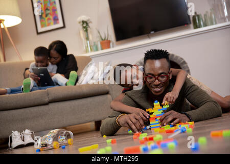 Father and son playing on the floor, mother and son reading on the couch Stock Photo