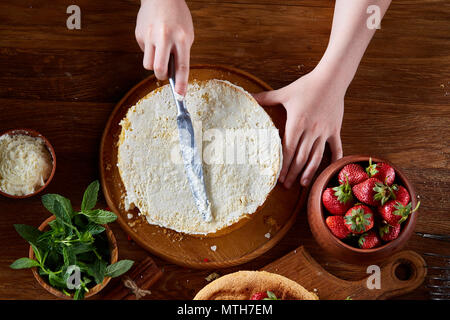 Close up of girl's hands adding some whipped sour cream on the top of delicious strawberry cake, selective focus. Tasty pastry is on the wooden table  Stock Photo