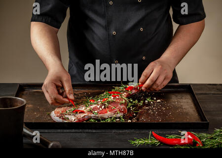 Male chef rubbing raw lamb shanks with greens and spices on stone tray on wooden table. Chef cooking appetizing shank of lamb. Stock Photo
