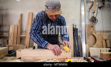 experienced carpenter in work clothes and small buiness owner working in woodwork workshop,  using sandpaper for polishing birdhouse at worktable in w Stock Photo