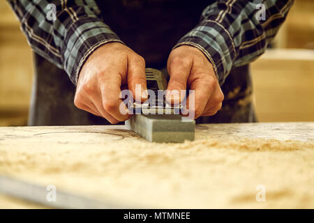 Close up of a experienced carpenter in work clothes and small buiness owner working in woodwork workshop, sharpening the tool with whetstone Stock Photo