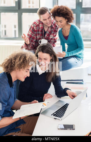 Two young students searching for an answer to their question both online and in a textbook while sitting at desk in the classroom during break Stock Photo