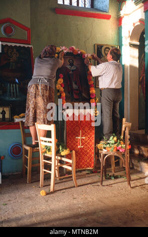 A couple decorate the icon with flowers in a rural church on the eve of the feast of St. Nikitas, Sellas, Peloponnese, Greece. Stock Photo