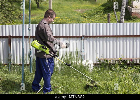 a man in glasses with a trimer mows the grass in the courtyard of a house in the village, brings order to the territory of the country Stock Photo