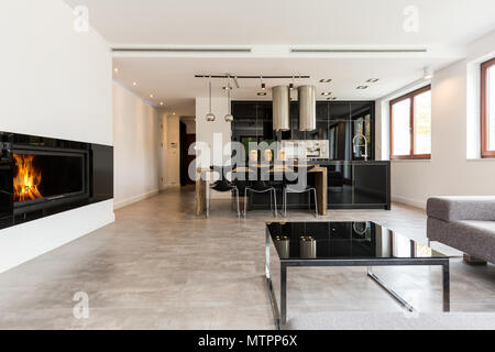 Spacious interior with fireplace, sofa, small table, dining set and black open kitchen Stock Photo